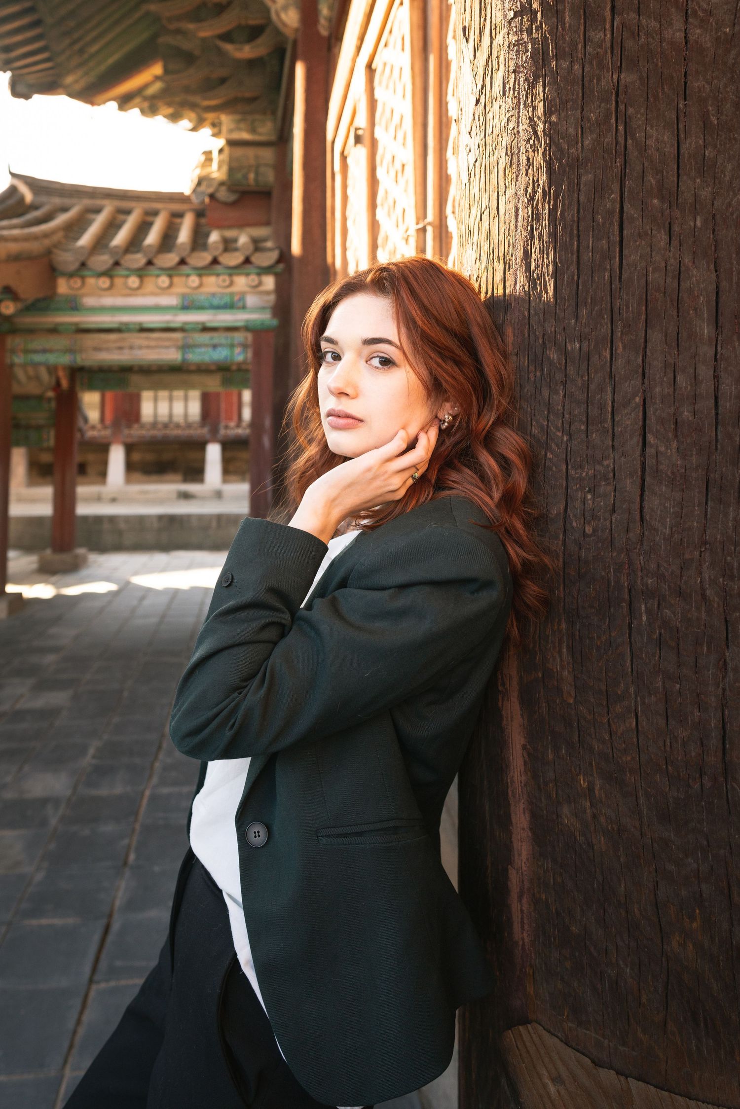 A woman in a black blazer poses thoughtfully against a wooden pillar at a traditional Asian temple.