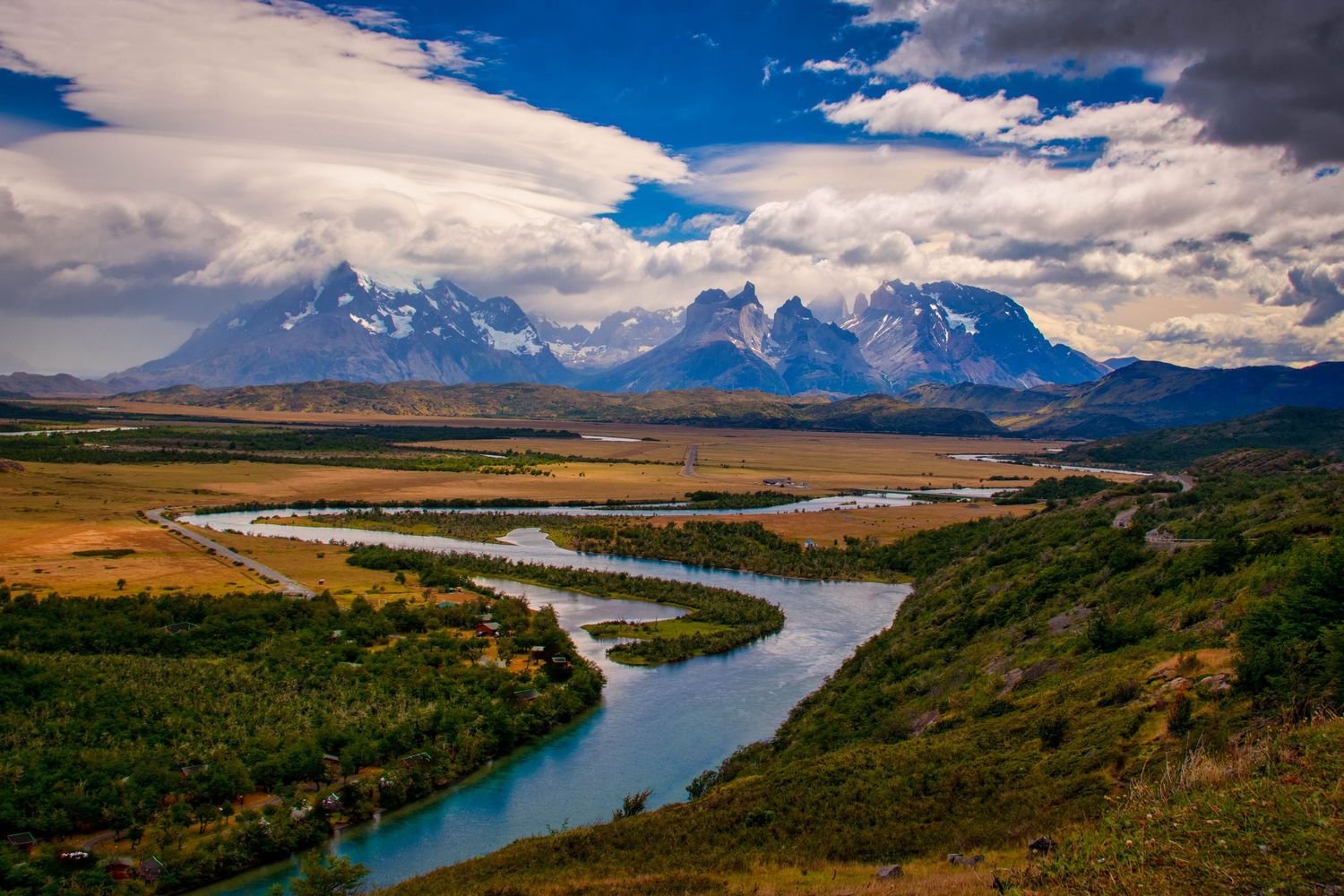 Panoramic view of winding river through rugged mountain landscape with dramatic clouds and golden grasslands.