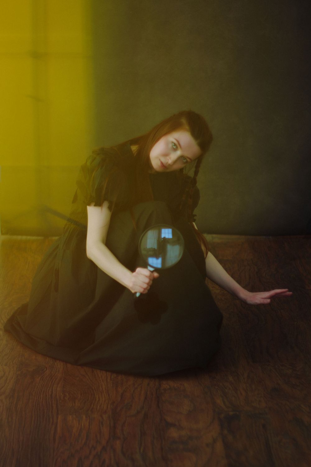 Young woman in black Victorian dress kneeling on wooden floor, holding magnifying glass with yellow light casting dramatic shadows.