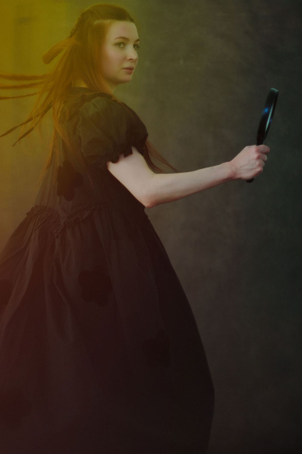 Woman in black Victorian dress holding a hand mirror, dramatic studio portrait with yellow and teal lighting.