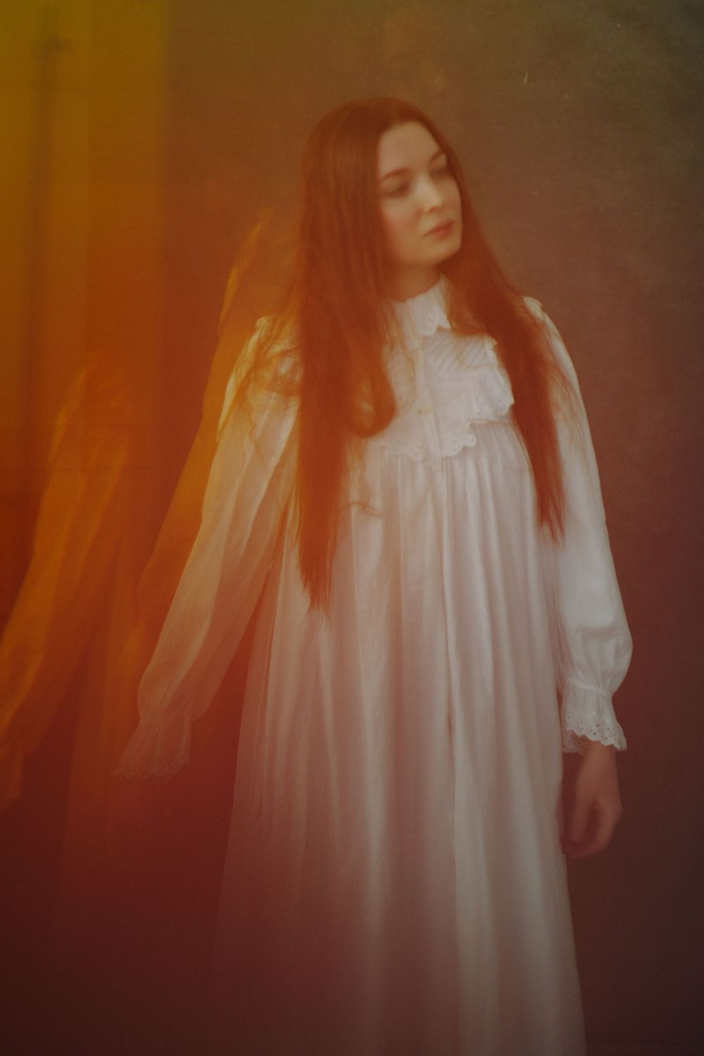 Young woman with long red hair in a white Victorian nightgown poses dramatically in warm orange atmospheric light.
