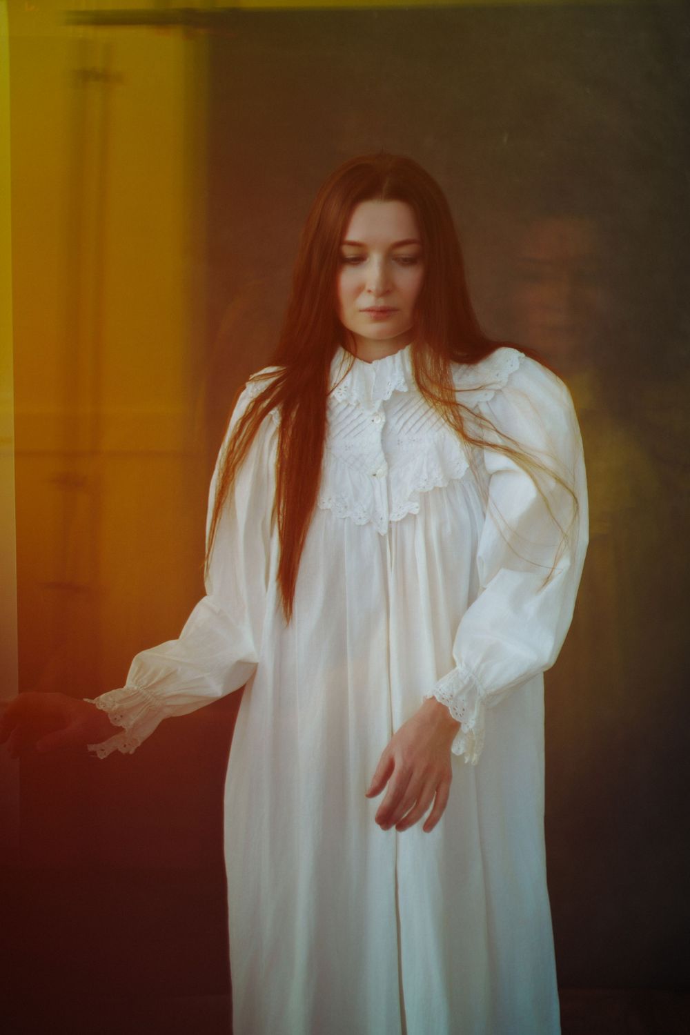 Woman in white Victorian nightgown with long red hair poses dramatically in warm golden light.