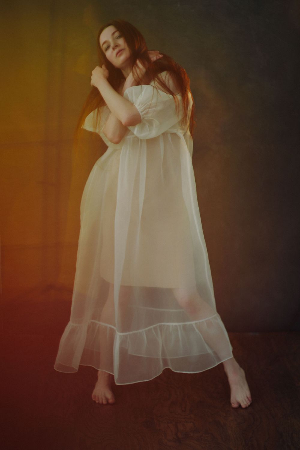 Woman in sheer white maxi dress poses barefoot against moody gradient background with warm amber lighting.