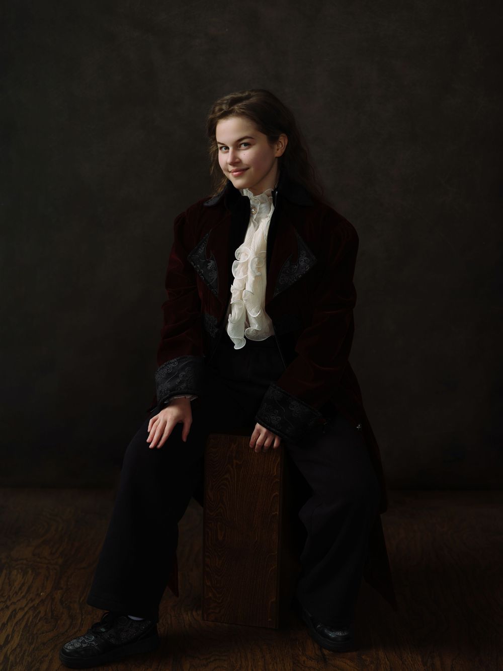 Young woman in vintage burgundy velvet coat and ruffled white shirt sits posed against dark studio backdrop.