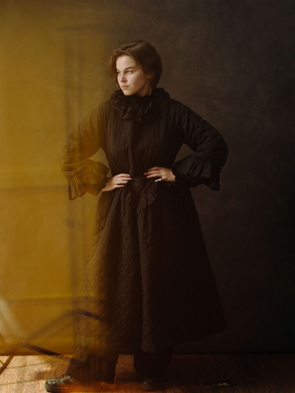 Woman in long black quilted Victorian dress poses confidently against golden draped background.