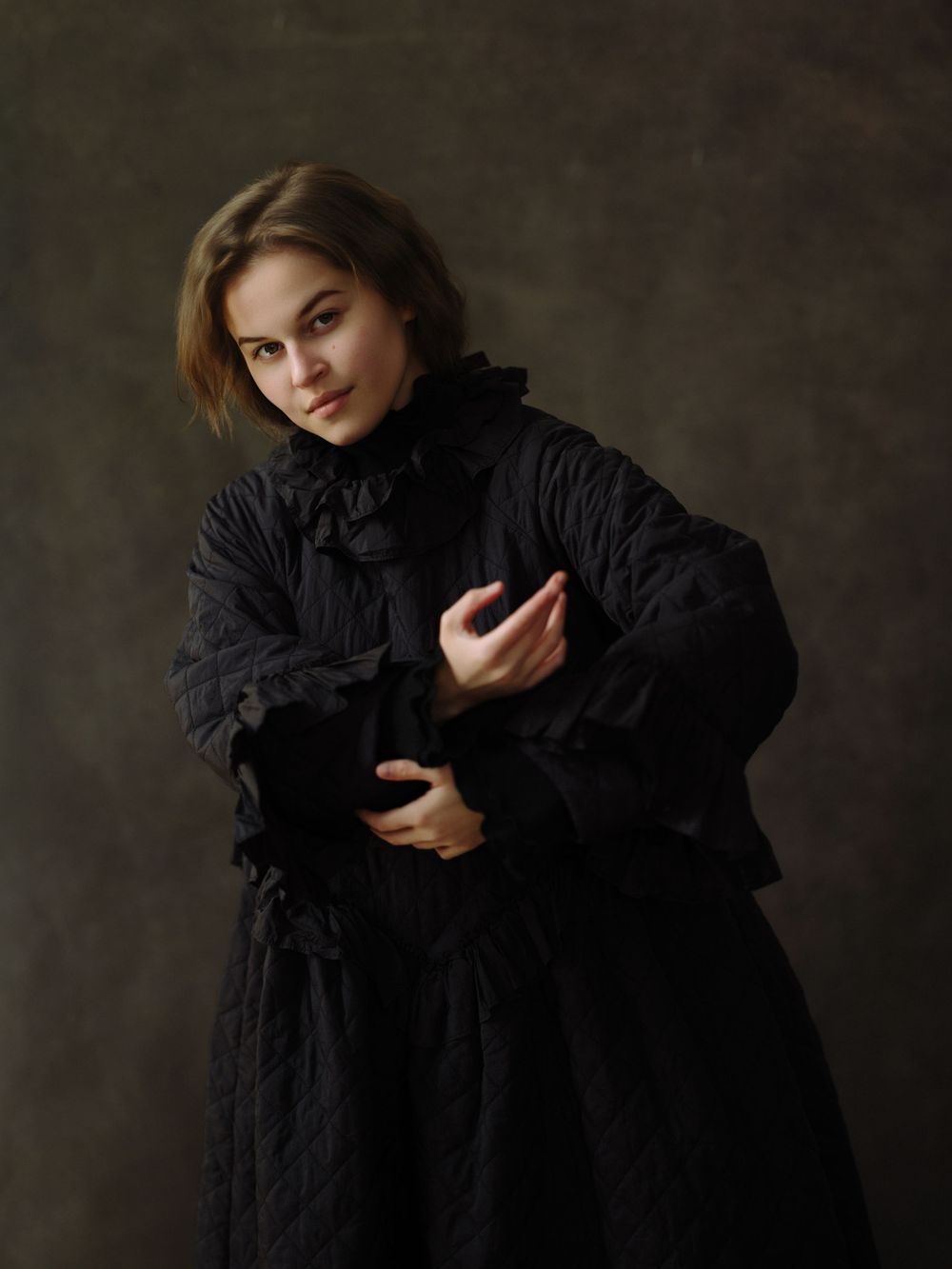 Young woman in black Victorian-style ruffled dress poses gracefully against dark studio background.