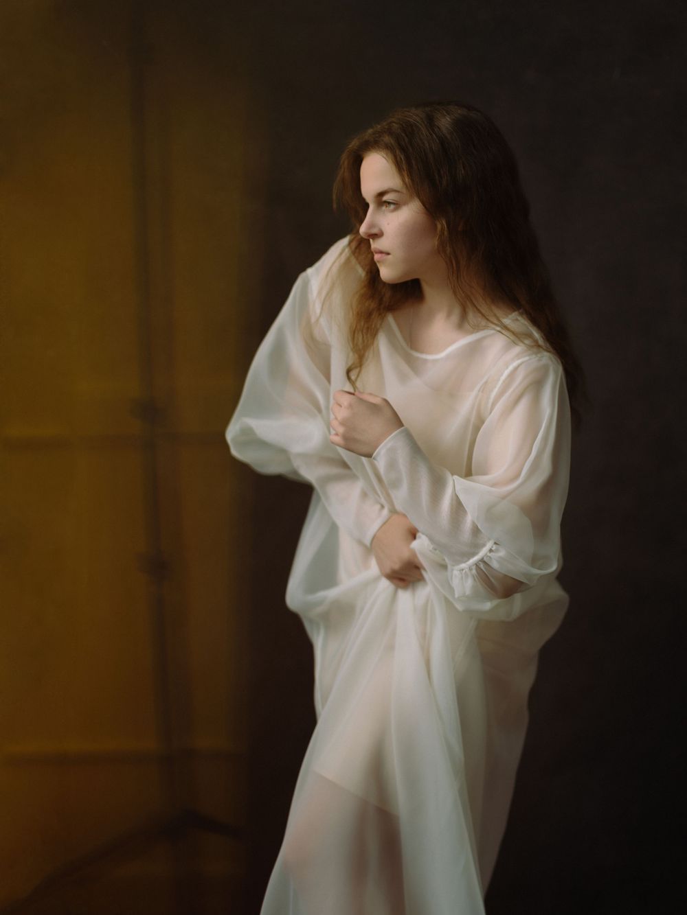 Young woman in sheer white oversized dress gazing sideways against dark dramatic background.