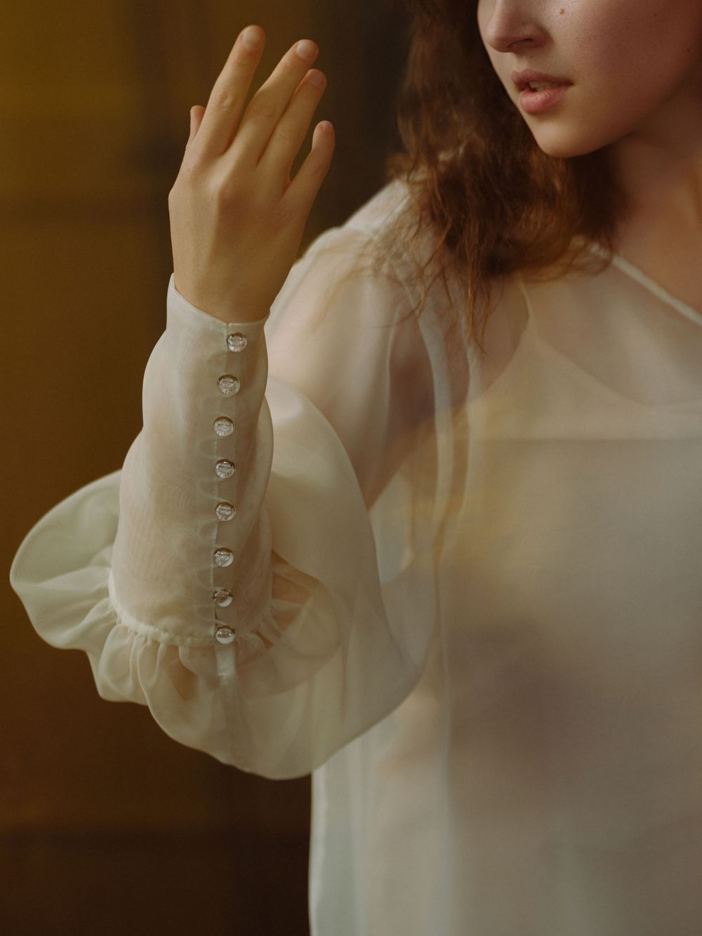 Woman in sheer ivory blouse with pearl button cuffs and puffed sleeves, raising her hand gracefully.