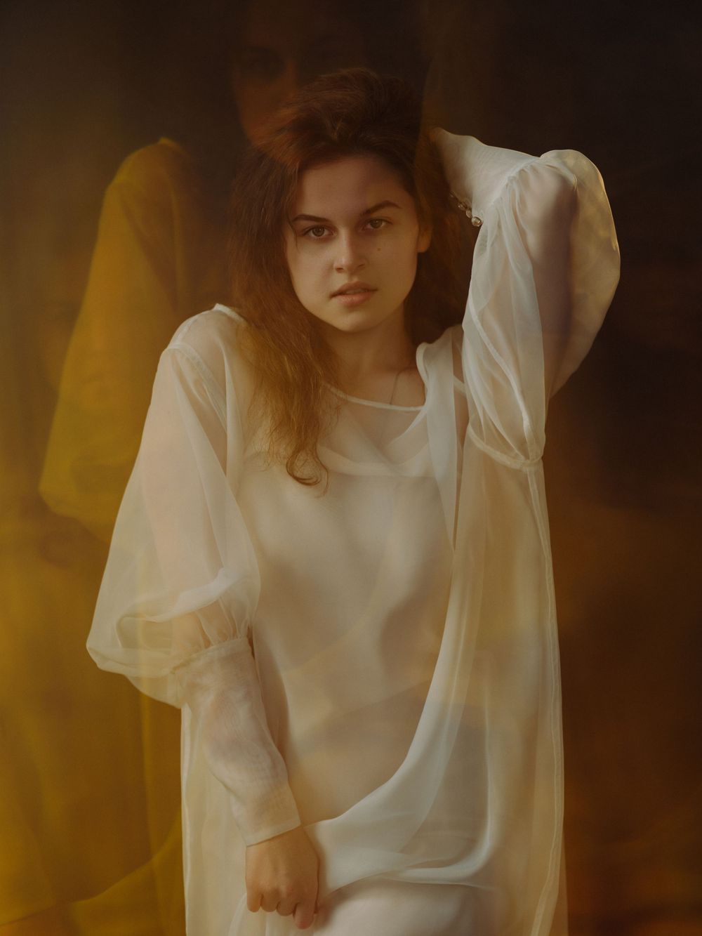 A young woman in a sheer white dress poses gracefully against a warm golden background.