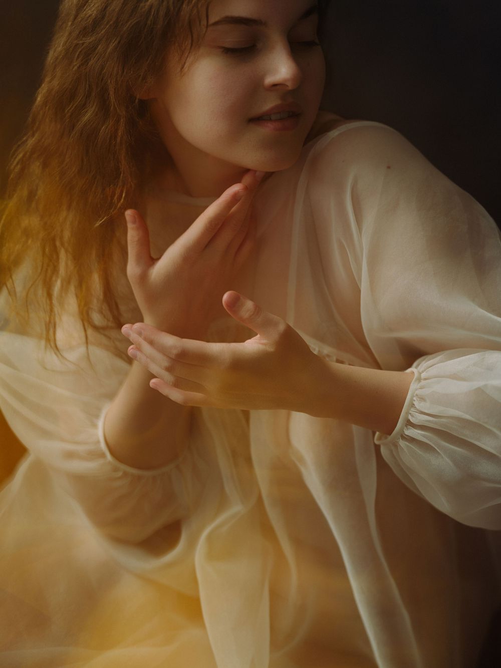 Young woman in white sheer dress with hands gracefully posed, soft golden light creating ethereal portrait photography.