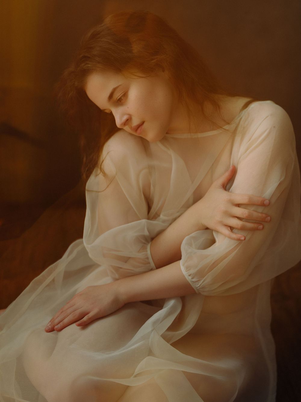 A young woman with auburn hair in a sheer white gown sits with eyes closed, arms crossed, in soft warm light.