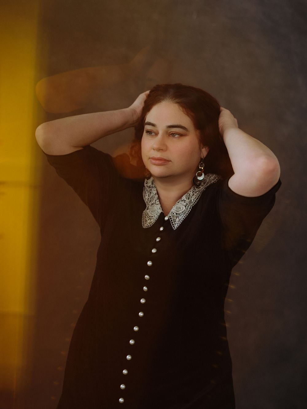 Young woman in black button-up dress with white collar poses with hands in hair against moody studio backdrop.