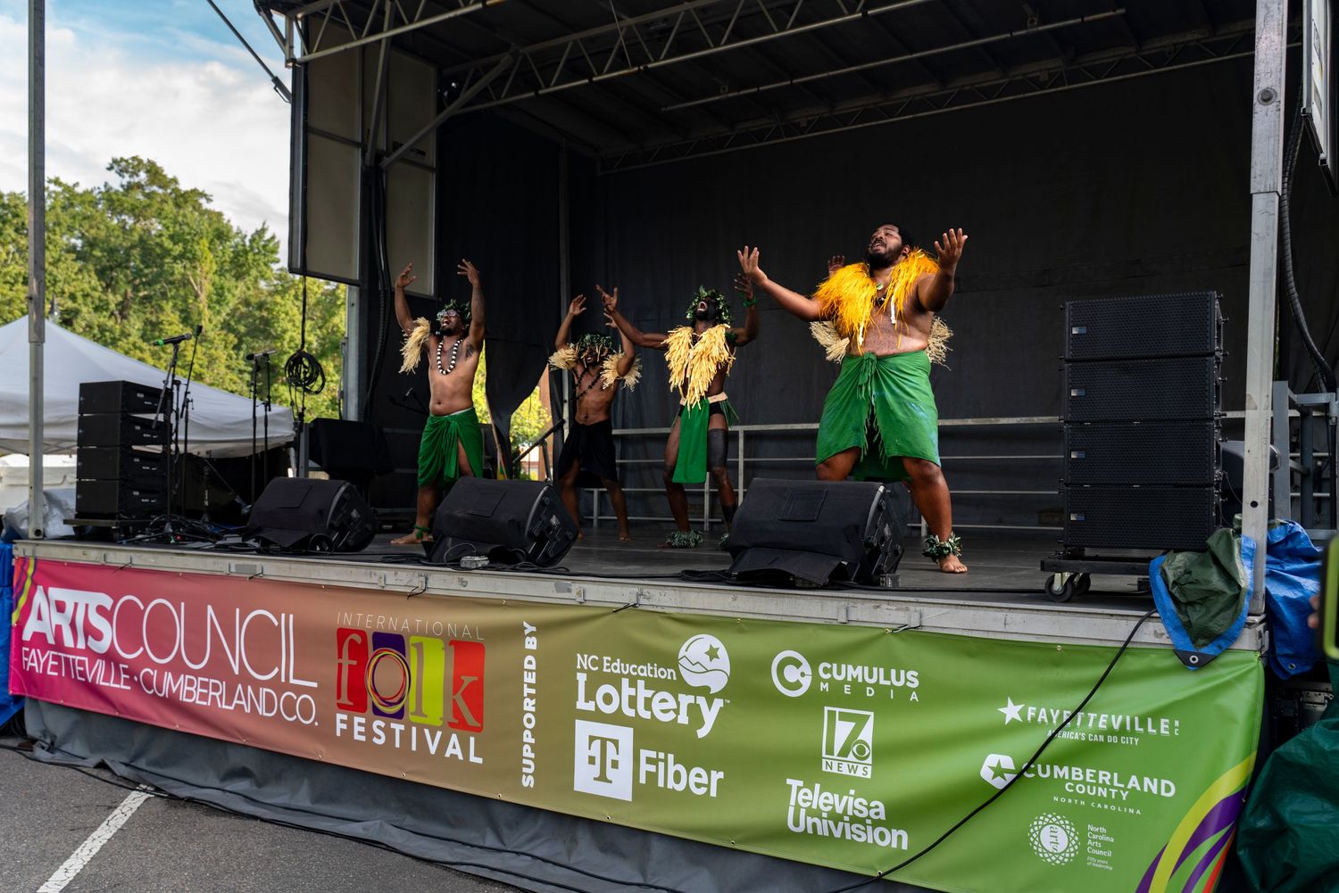 Performers in yellow and green outfits dance energetically on an outdoor festival stage with sponsor banners below.