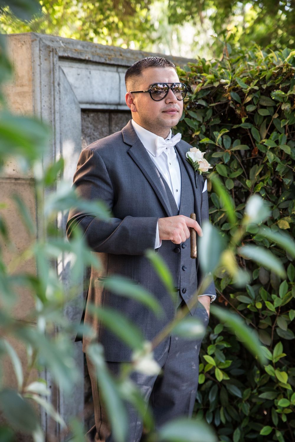 A person in a black suit and sunglasses stands among green foliage in an outdoor garden setting.