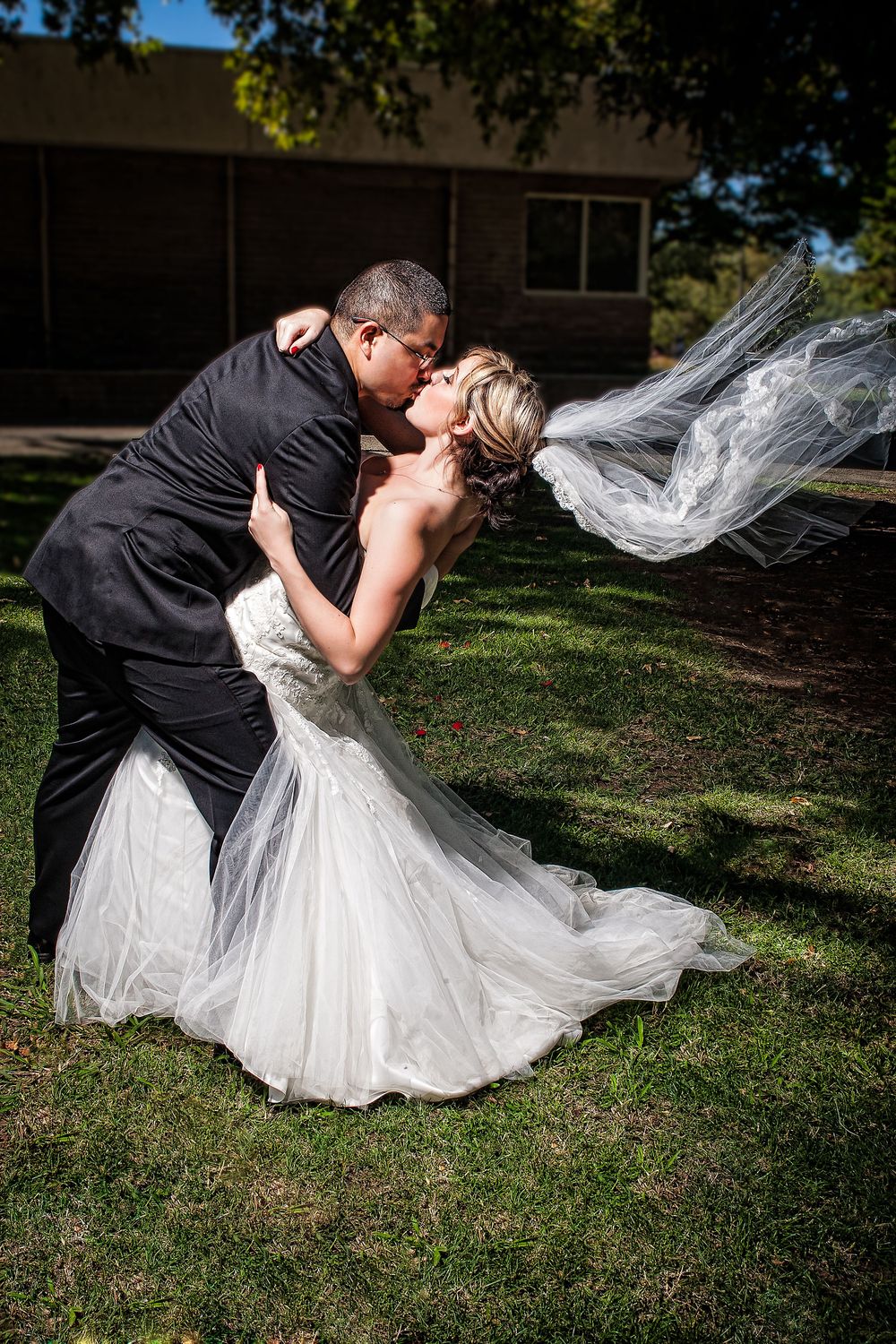 A romantic wedding couple shares a dip kiss on a grassy lawn with the bride's veil flowing in the breeze.
