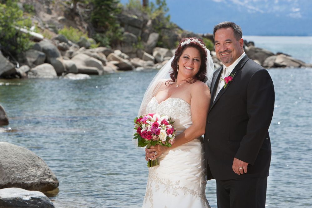 A couple in wedding attire poses on a rocky beach at Lake Tahoe with bright blue water and mountains in background.