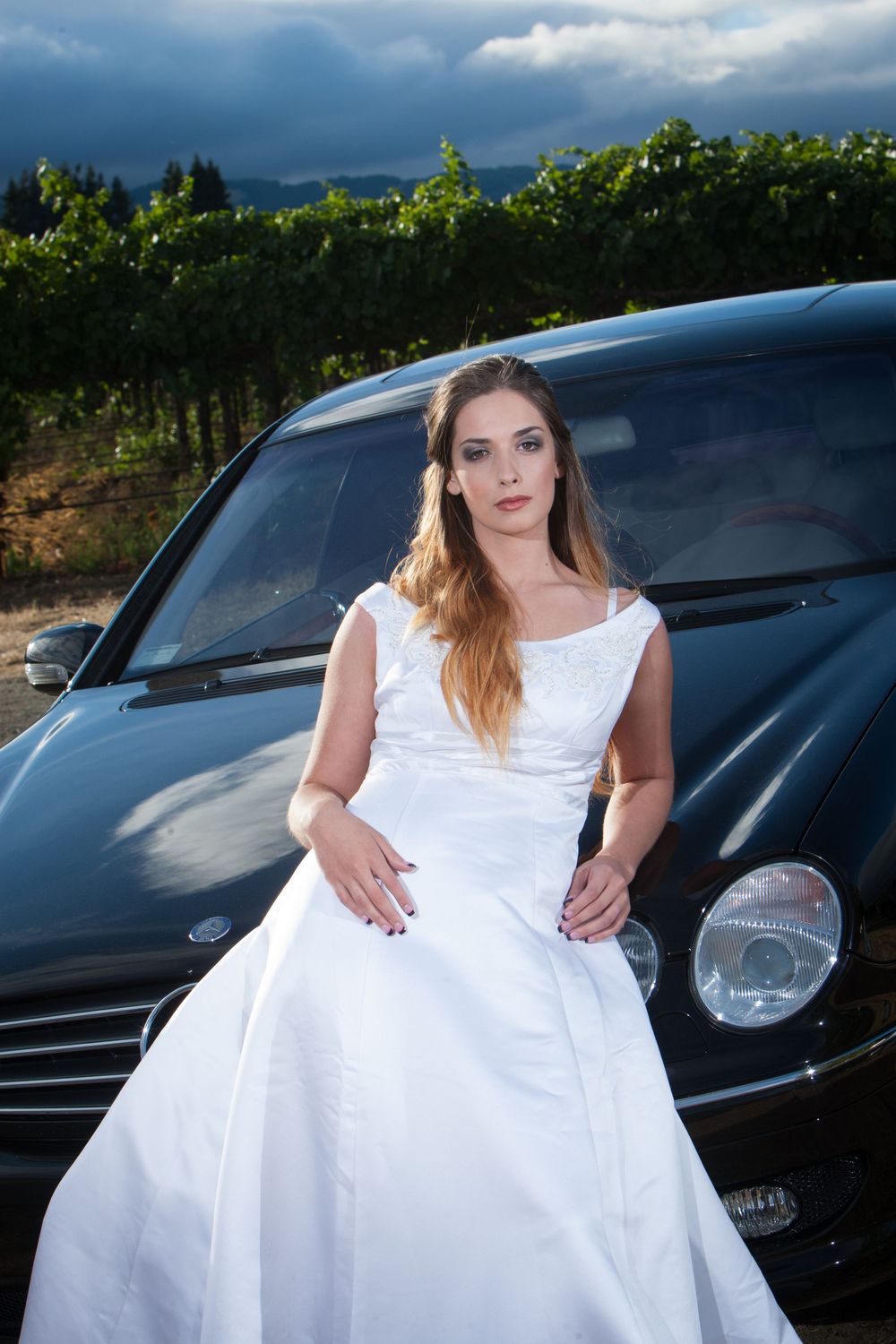 A bride in a flowing white wedding dress poses against dark vehicles and vineyard scenery at sunset.