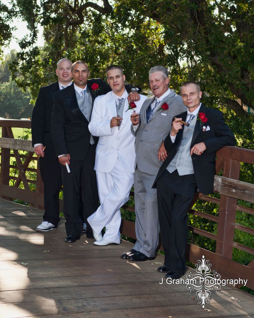 Groomsmen in formal attire pose casually on a wooden bridge during an outdoor wedding photo session.
