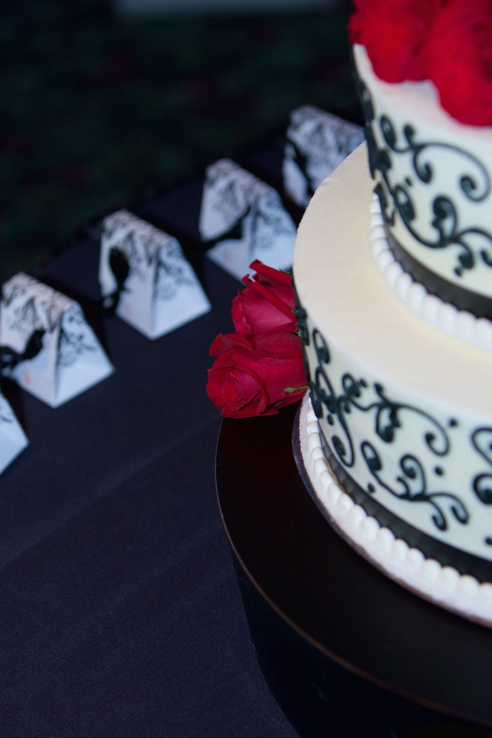 Elegant white wedding cake decorated with black filigree designs and red roses, with matching favor boxes in background.