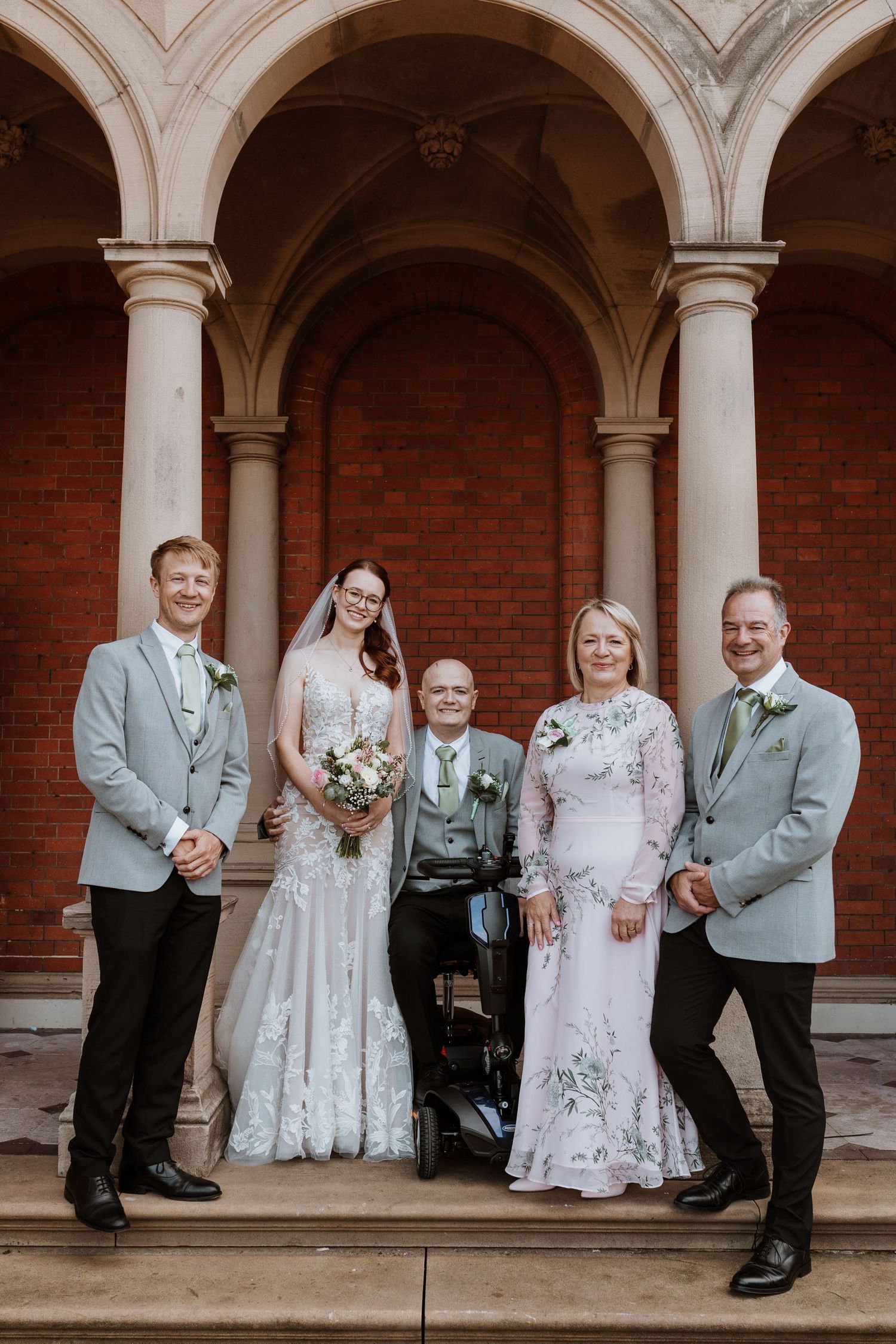 A wedding party sits together on stone steps against a brick wall with white arched columns.
