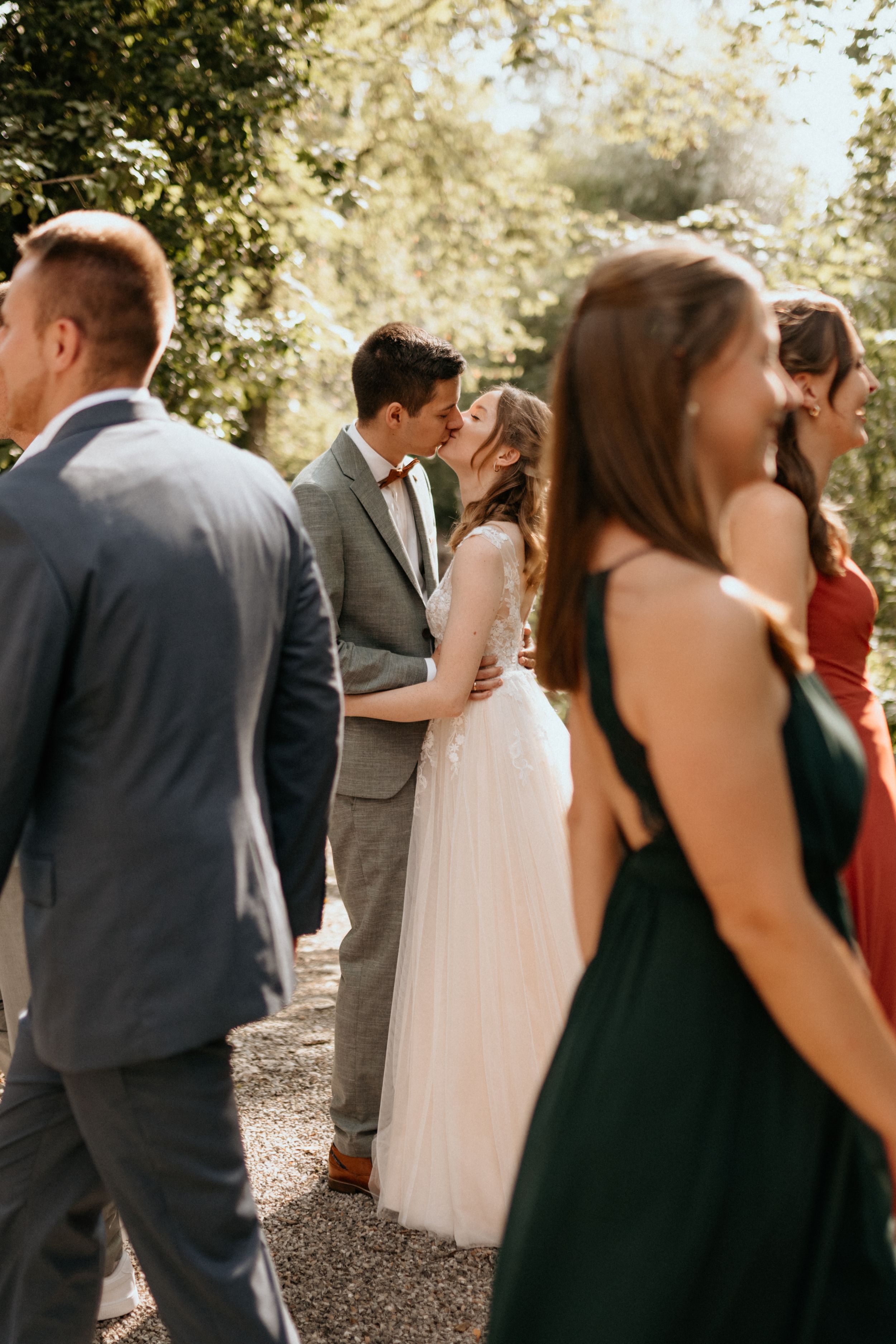 Wedding party members in dark dresses and gray suits walking together on a sunny outdoor path.