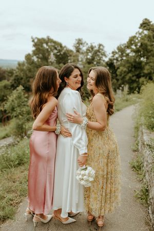 Three friends in pastel dresses sharing a tender moment on a countryside path.
