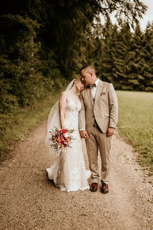 Newlyweds walking together on a rustic dirt path holding a red bouquet.