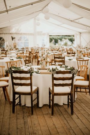 Wedding reception venue with white tablecloths and wooden chairs set up under an elegant white tent.