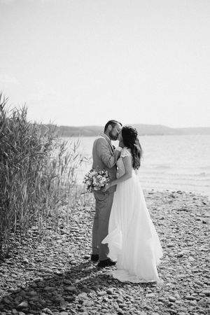 Black and white beach wedding photo with couple embracing on rocky shoreline.
