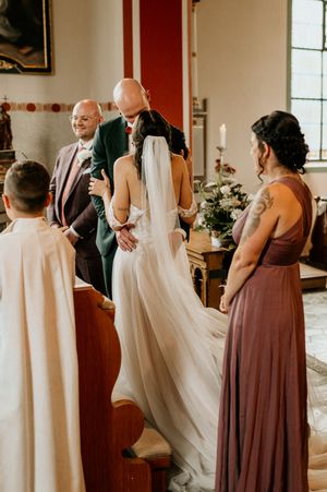 Wedding party members in burgundy dresses helping with the bride's gown in church.