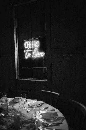 Dark reception room with glowing neon signs reading 'Cheers' and 'To Love'.