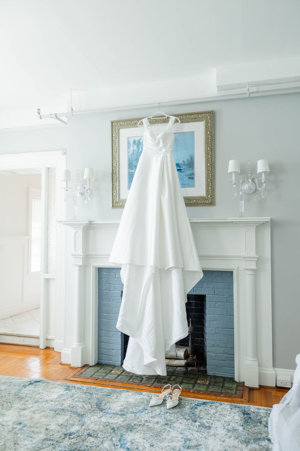 White wedding dress displayed in front of classic blue fireplace with white mantle and hardwood floors.