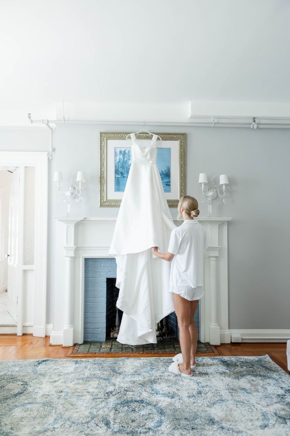 A white wedding gown hangs against a fireplace mantel in an elegant room with blue walls and area rug.