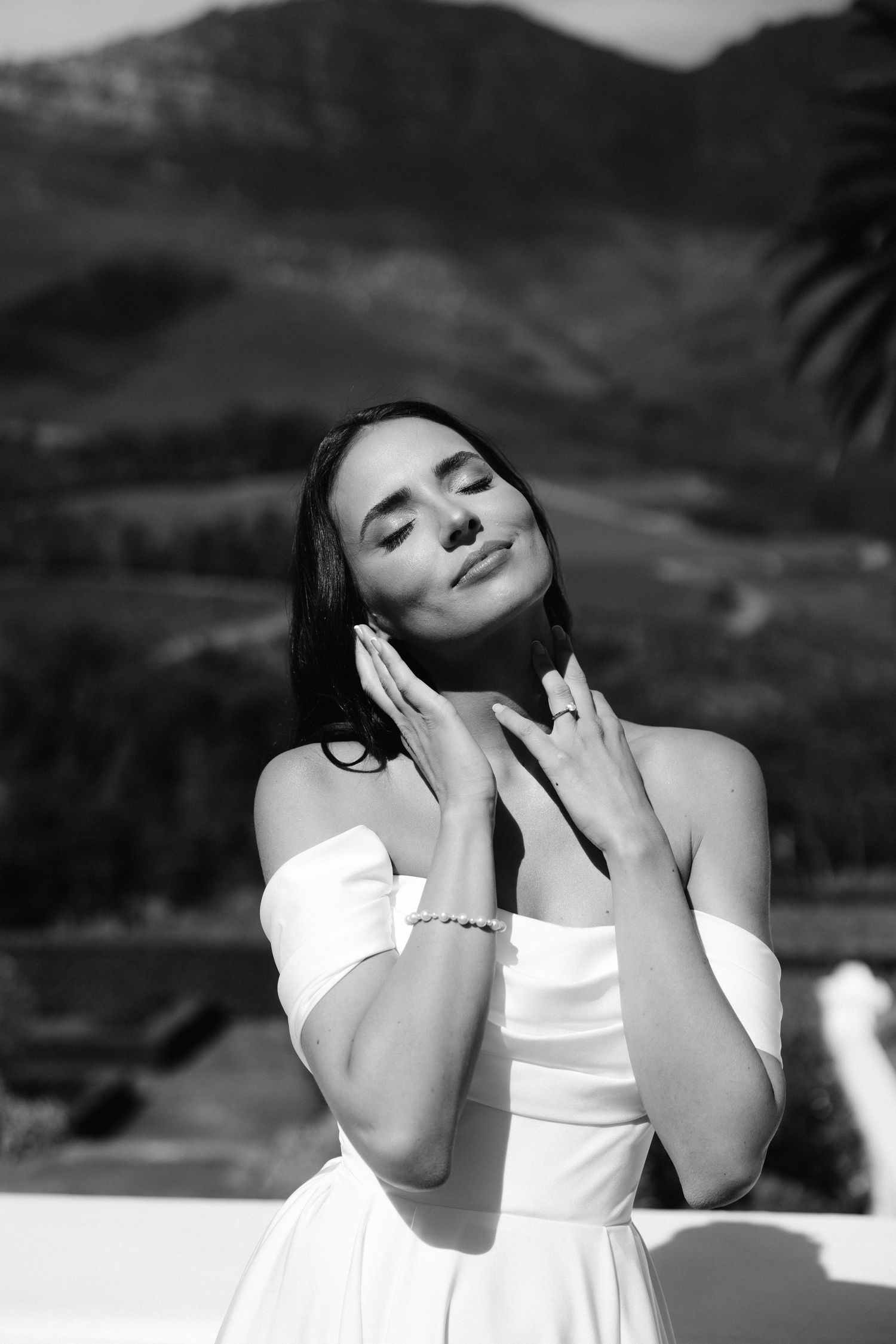 A person in a white dress poses gracefully against a mountain landscape backdrop in black and white photography.
