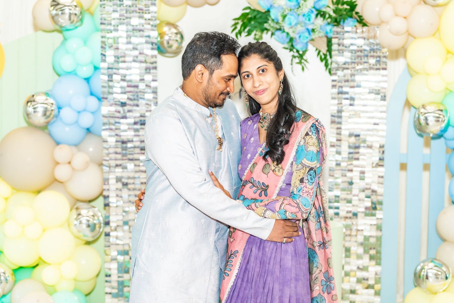 A couple in traditional Indian attire embraces at a celebration with pastel balloon decorations and floral accents.