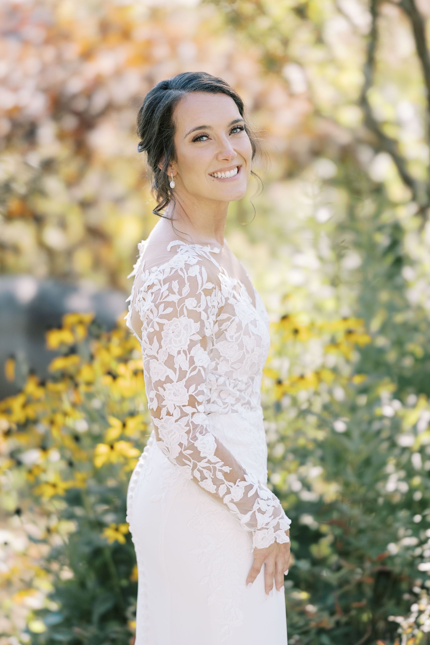 A person in a white lace wedding dress poses outdoors with yellow flowers and autumn foliage in the background.