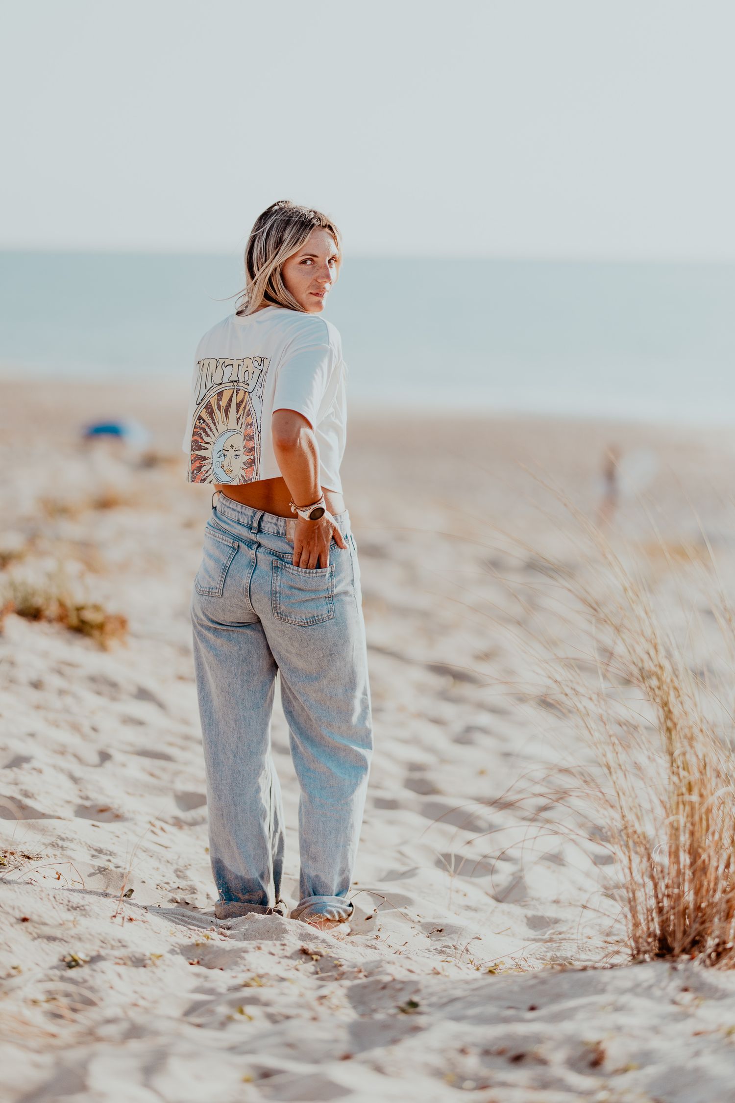 Une personne portant un jean délavé léger et un haut marron se tient sur une plage de sable près de l'herbe séchée au coucher du soleil.