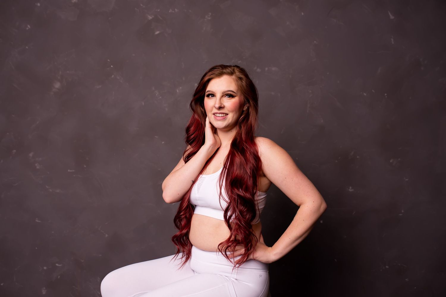 Woman with long red wavy hair posing confidently in a white crop top and pants on gray backdrop.