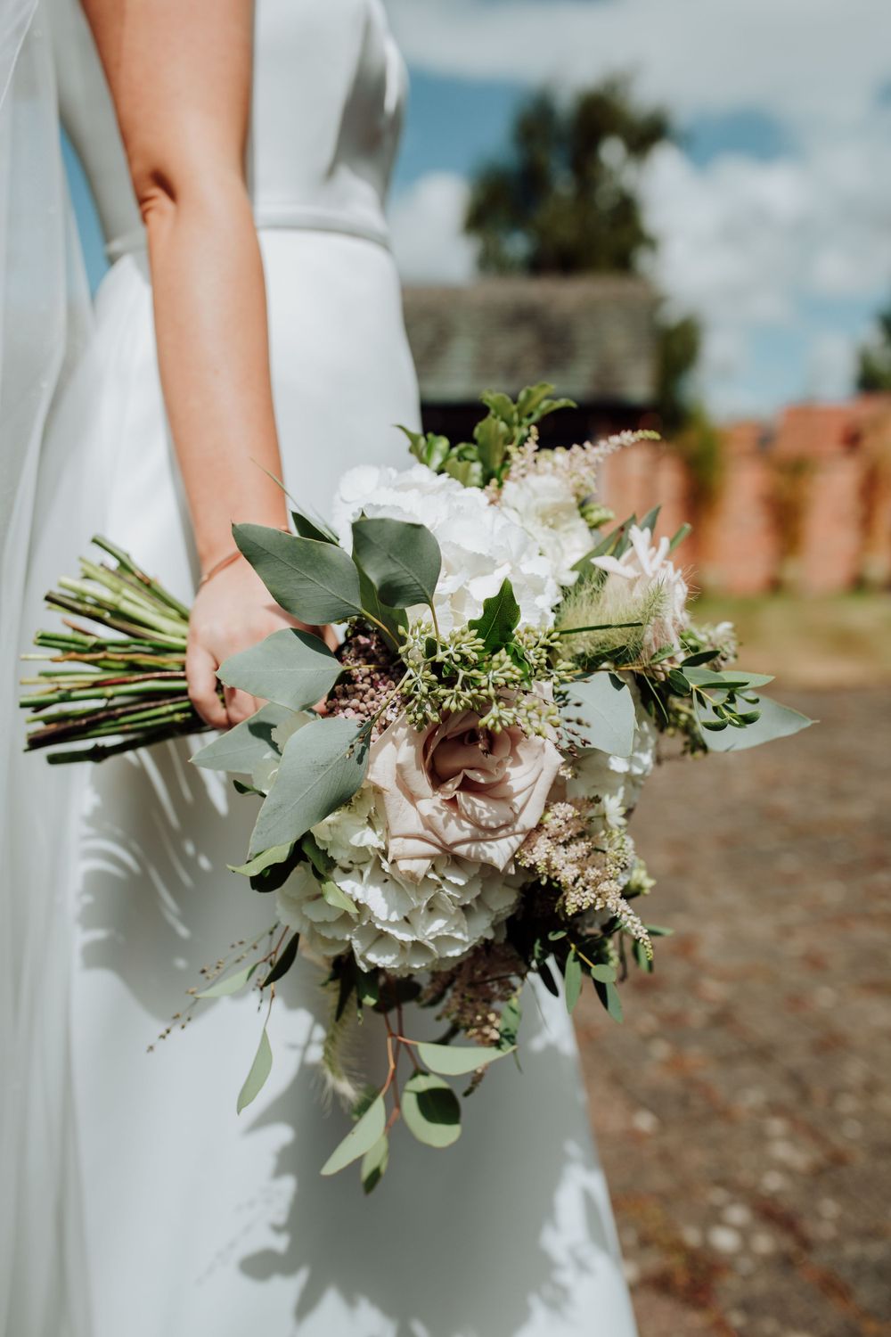 A bride in a white dress holds an elegant bouquet with blush roses, white flowers and eucalyptus leaves outdoors.