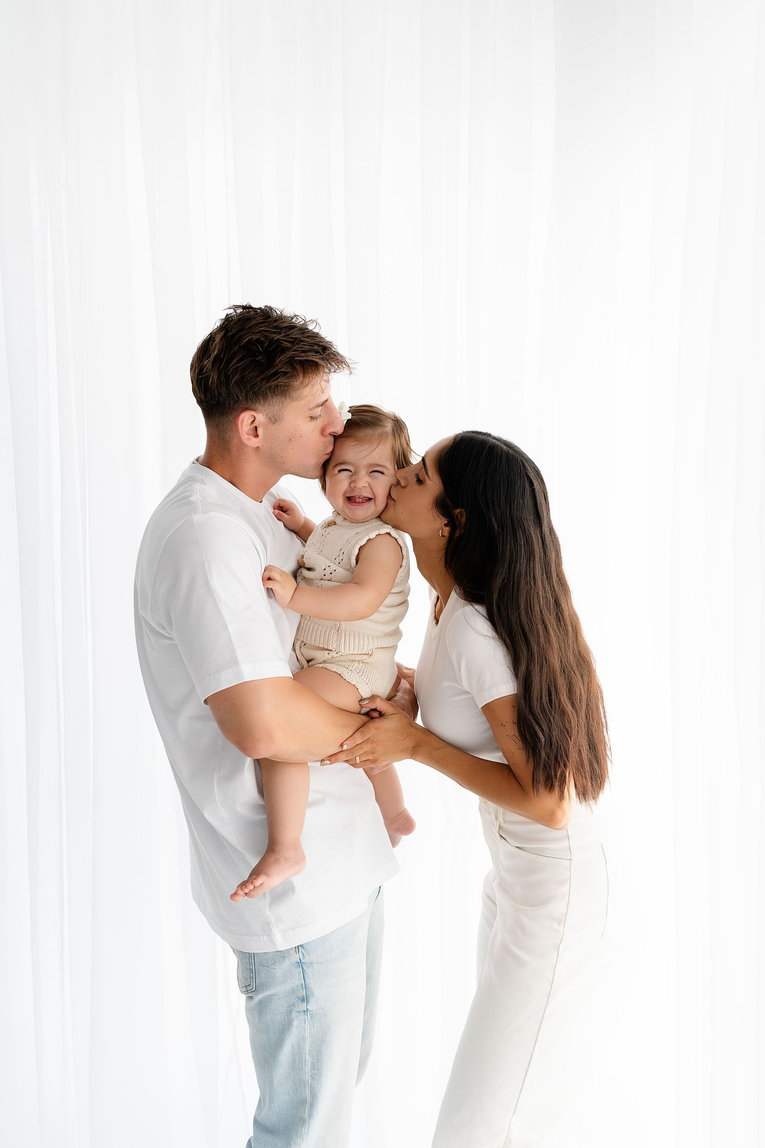 Family of three shares intimate moment against white background in matching light outfits.