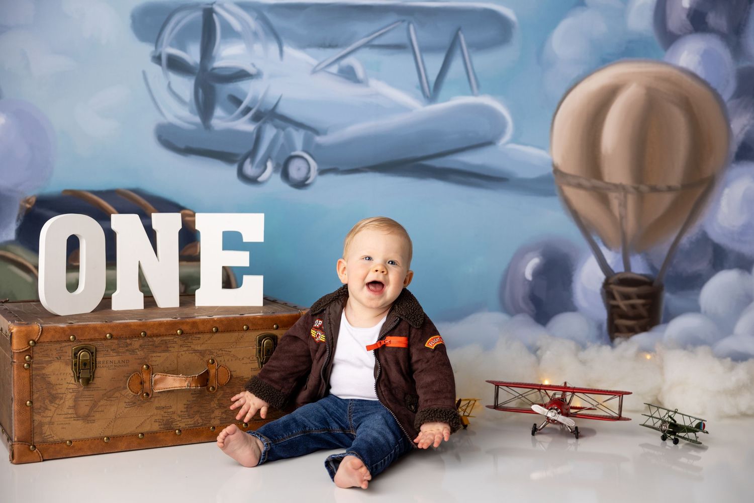 Baby in brown jacket sitting by vintage suitcase with vintage aviation themed backdrop featuring planes and hot air balloon.