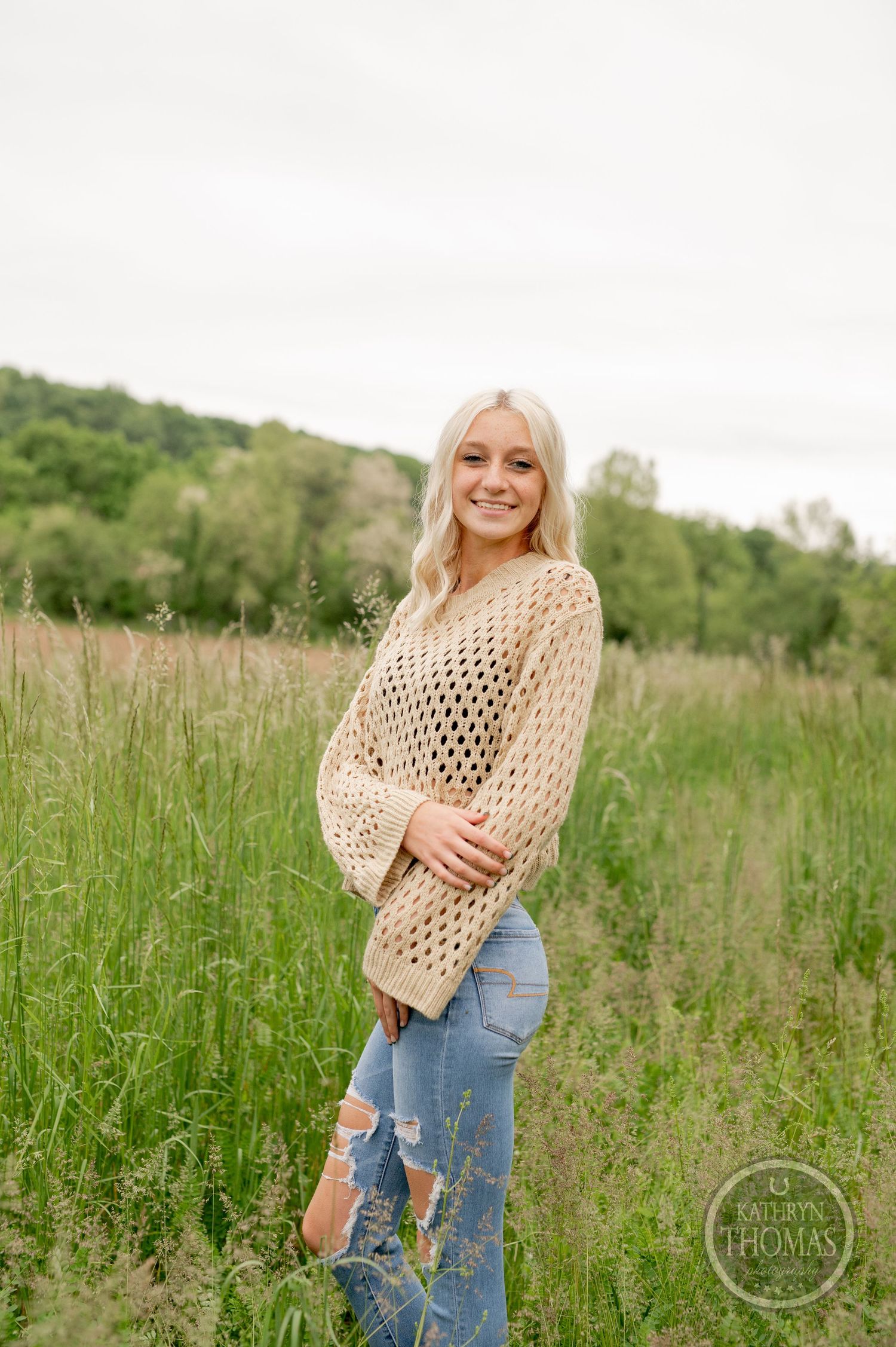 Relaxed outdoor portrait showcasing a chunky knit beige sweater and distressed blue jeans in tall grass.