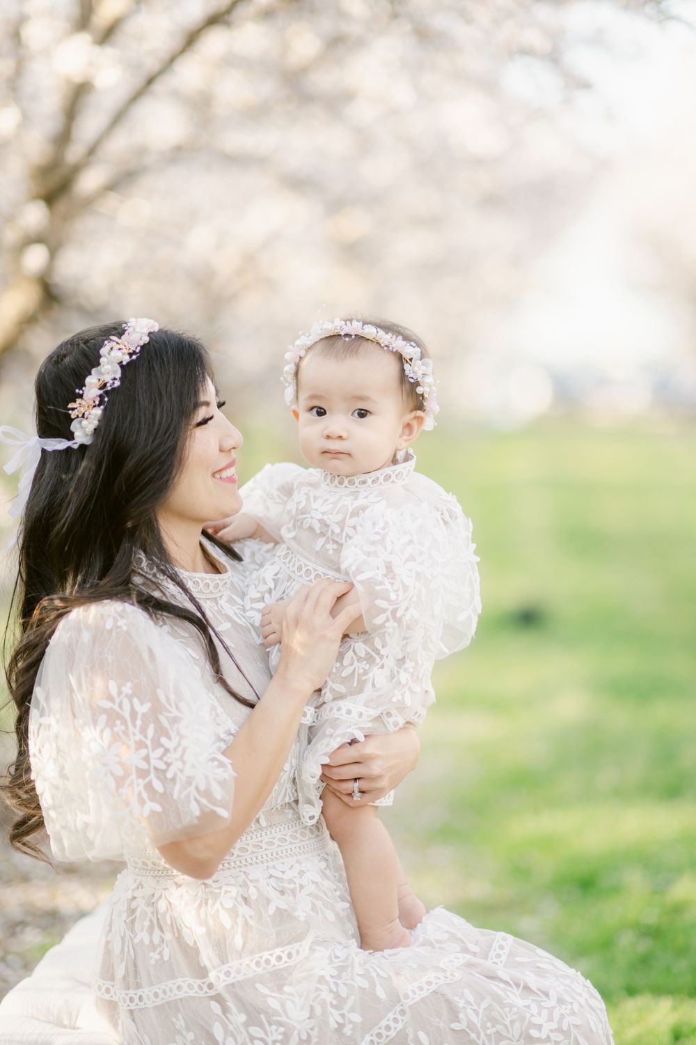 A parent and baby wearing matching white lace dresses pose under cherry blossoms.