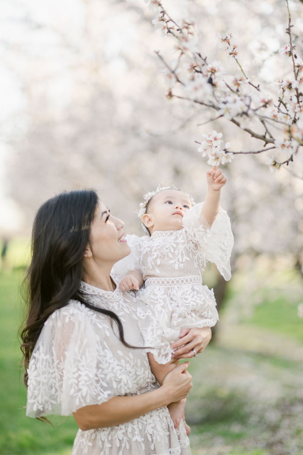 A parent holds up a baby in a white dress to touch cherry blossom branches.