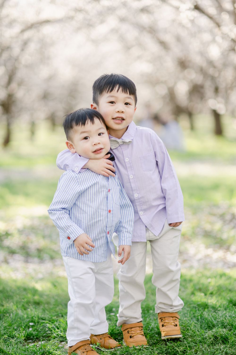Two small children in matching button-down shirts and white pants pose together under blooming cherry blossom trees.