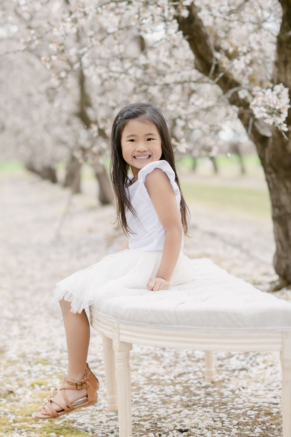 A young person in a white dress sitting on a white bench under blooming cherry blossom trees.