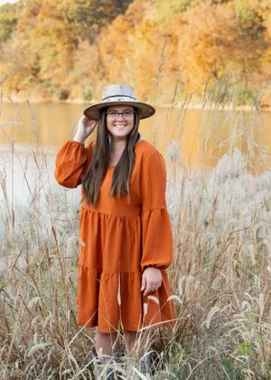 Person in orange dress and sun hat standing among tall grass by autumn lake.