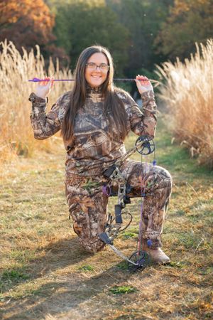 Archer in camouflage gear posing with compound bow in golden fall setting.