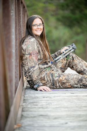 Hunter in camouflage gear resting on wooden bridge with equipment.