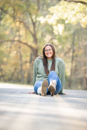 A student sits on a road wearing a mint green sweater, blue jeans and glasses in an autumn setting.